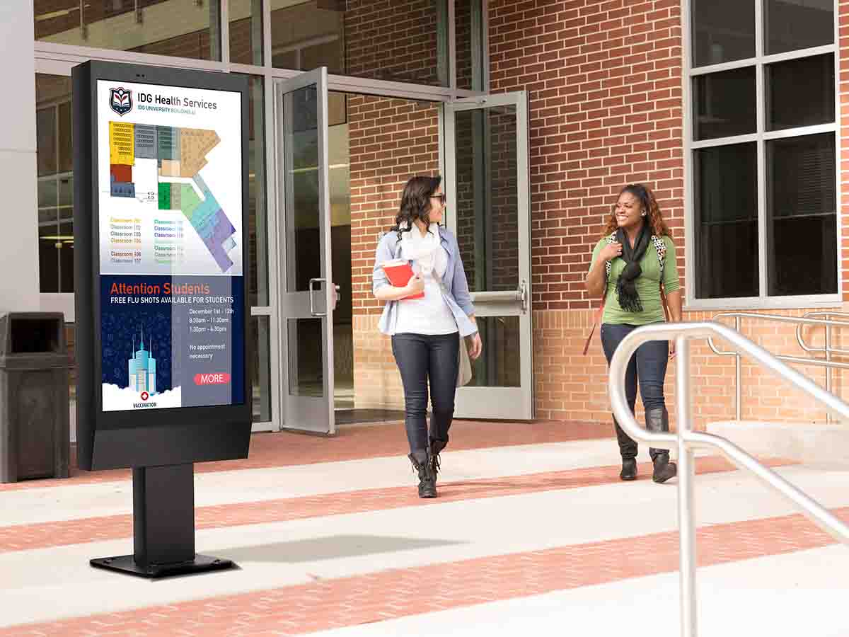 Two female students walking outside a school and past digital signage.